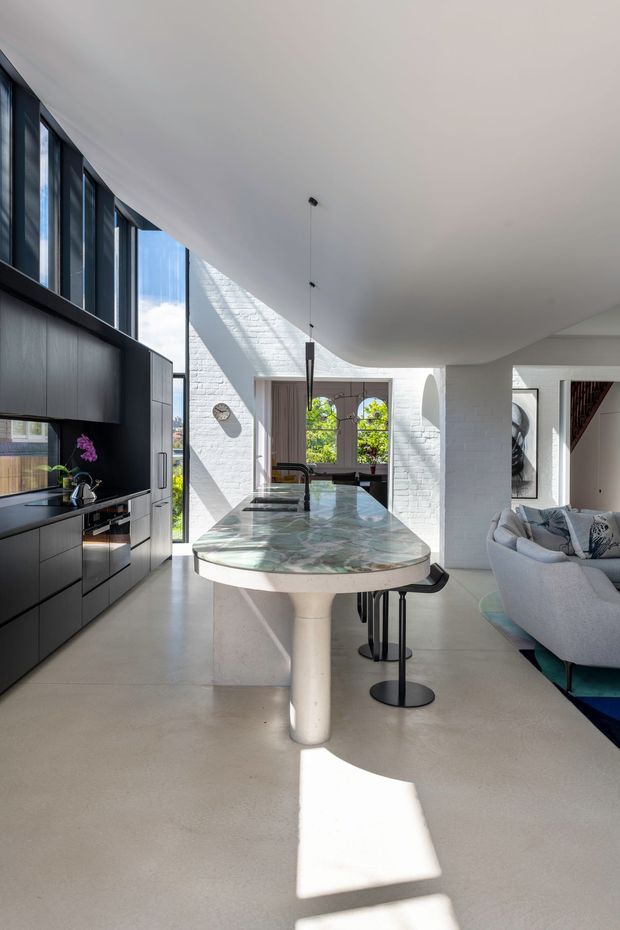 Bright open-plan kitchen and living area with a sculptural curved island featuring a green-and-white stone benchtop, dark built-in cabinetry, tall black-framed glazing, white painted brick walls, and a smooth light floor. A partially visible lounge sits to the right, while strong sunlight creates dramatic geometric shadows across the room.