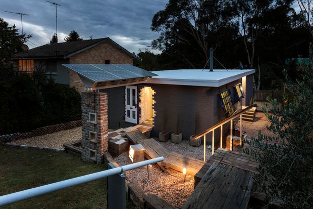 A dusk exterior view of a compact backyard structure with a flat metal roof, dark cladding, exposed brick and timber elements, solar panels over a covered entry, and raised timber decking with steps. The building sits within a leafy suburban garden, with a larger brick house visible behind it.