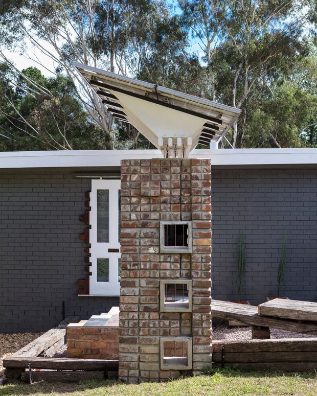 Exterior view of a low, contemporary brick building in a wooded setting. A dark charcoal brick wall sits beneath a flat roof, with a white door and a prominent freestanding brick chimney or outdoor fireplace structure in front. The chimney has multiple small openings and a broad angled metal canopy with exposed rafters. Rustic timber retaining elements and sparse planting occupy the foreground.
