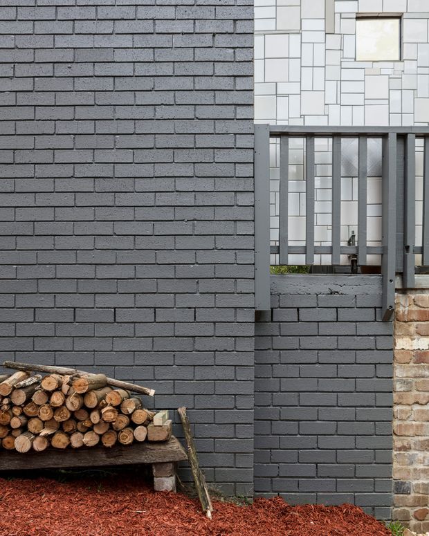 Close-up exterior architectural view showing a dark grey painted brick wall with a recessed opening and grey metal balustrade. Behind it is a contrasting white tiled or masonry facade with a small square window. A weathered timber bench stacked with firewood sits at the base, bordered by red mulch and a partial exposed brick wall at right.