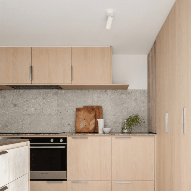 A compact contemporary kitchen with extensive light timber cabinetry, integrated upper cupboards, a black built-in oven and cooktop, and a grey stone-look tiled splashback. A slim timber-toned rangehood shelf, dark benchtop, cutting board, bowl, and small plant add functional and decorative details. The composition focuses on the kitchen joinery and work area.