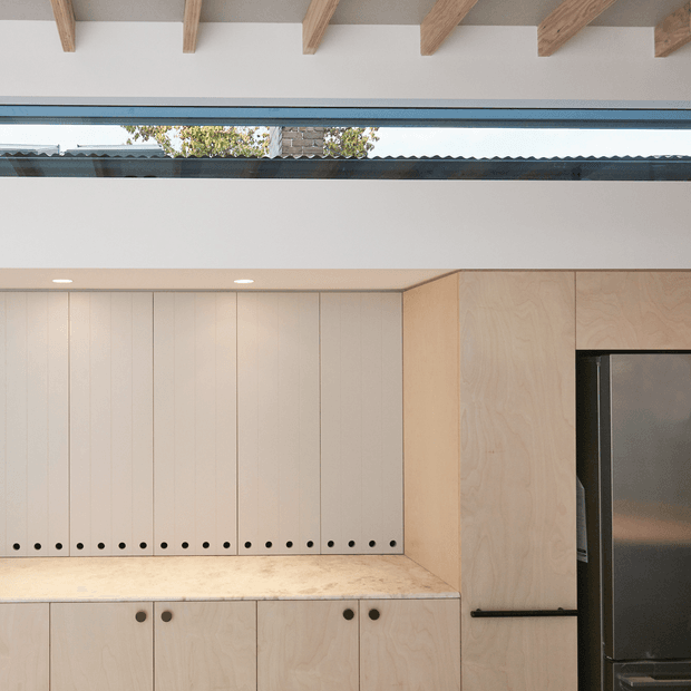 A minimalist built-in kitchen with pale timber cabinetry, a light stone benchtop, integrated black hardware, and a tall dark refrigerator at right. The upper cabinetry features vertical panel detailing and small round ventilation or peg-like elements along the base. Exposed timber ceiling beams and a high horizontal clerestory window bring natural light into the space.