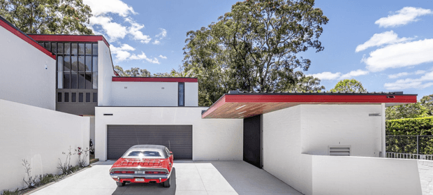 A contemporary white rendered residential building viewed from the driveway, featuring crisp geometric volumes, a low-pitched flat roof with vivid red metal edging, tall glazed stairwell windows, and an integrated double garage. A red classic car is parked on the pale concrete driveway. Mature trees and blue sky form the background, while a partially screened side passage and raised terrace wall are visible to the right.