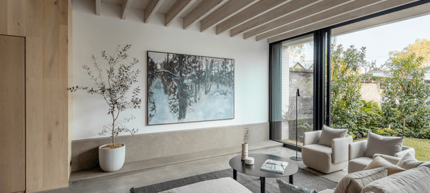 A refined contemporary living room with a neutral palette, expansive black-framed glazing, exposed light timber ceiling beams, built-in timber wall joinery, a long stone or concrete bench, sculptural artwork, upholstered lounge chairs and a round black coffee table. The room opens visually to a leafy garden and receives abundant natural light.