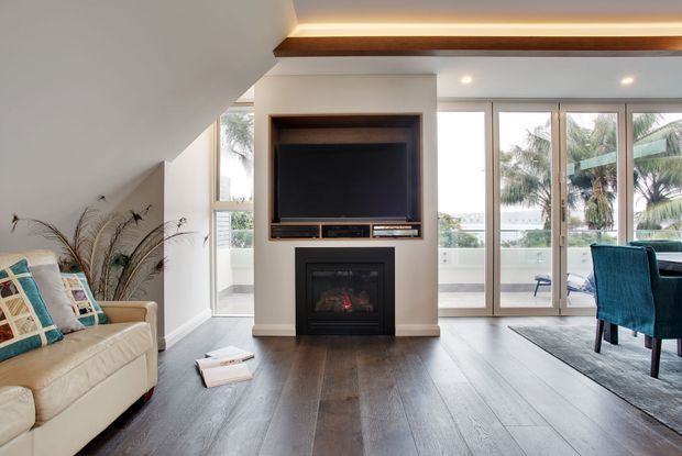 Bright contemporary living room beneath a sloped ceiling, featuring dark timber flooring, a built-in television and fireplace wall, recessed display joinery, a cream sofa with patterned cushions, and full-height glazed doors opening toward a balcony or exterior view.