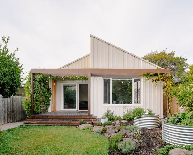 Front exterior view of a compact contemporary single-storey dwelling with a steep asymmetrical gable roof, pale vertical panel cladding, and a timber-framed covered porch. Large glazed doors and windows open onto a timber deck, with climbing greenery, raised corrugated metal garden beds, and landscaped planting around the facade.