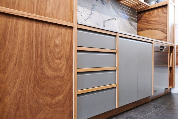 Close-up view of a contemporary kitchen or compact utility fit-out, featuring warm timber cabinetry, matte grey drawer and door fronts, a veined stone splashback or wall panel, open timber shelving, and dark tiled flooring. The image focuses on fixed joinery and cabinetry details.