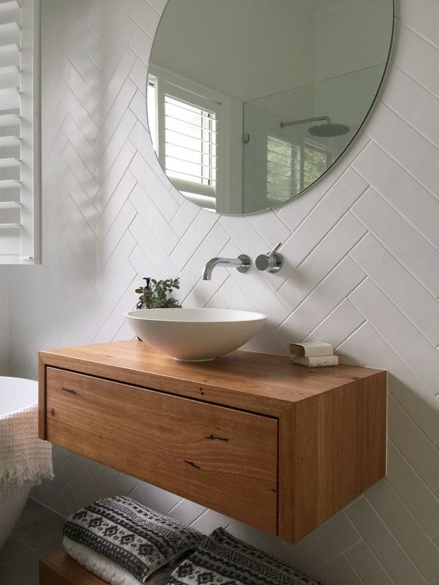 A bright, compact bathroom featuring a wall-mounted timber vanity with a white vessel basin, brushed metal wall-mounted tapware, and a large rounded mirror. The walls are clad in white diagonal herringbone tiles, with a louvred window and subtle greenery. A patterned upholstered stool sits beneath the vanity.