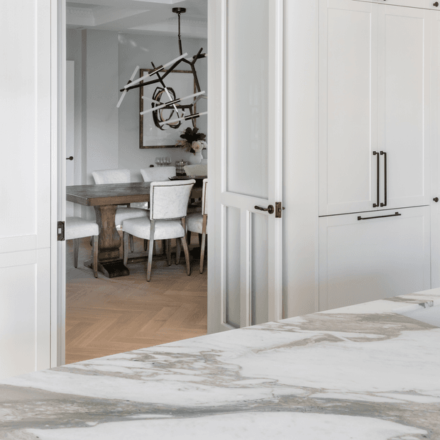 View from a bright white kitchen through glazed double doors into a refined dining room. The foreground features a large veined stone island benchtop and tall white built-in cabinetry with dark metal handles. Beyond, a timber dining table with upholstered chairs sits on a herringbone timber floor, beneath a sculptural branch-like pendant light and abstract wall art.
