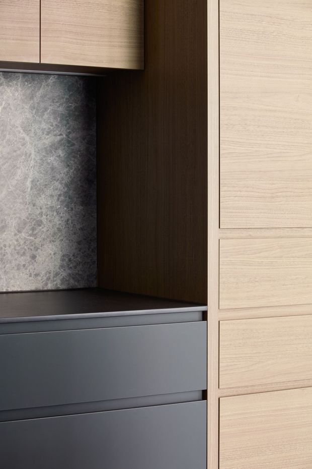 Close-up view of contemporary built-in cabinetry and benchtop joinery. Light timber-grain upper and tall drawer fronts contrast with dark brown vertical timber cabinetry and matte dark grey drawers. A grey veined stone splashback or feature wall sits behind a dark benchtop, with crisp minimal detailing and handleless fronts.