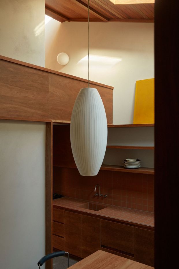 Warmly lit, compact kitchen or kitchenette with dark timber cabinetry, open shelving, a terracotta-red tiled benchtop and backsplash, and a small sink. A large white ribbed pendant hangs centrally beneath a timber-lined ceiling with a skylight. The space has cream plaster walls, strong mid-century-inspired timber detailing, and a bold yellow artwork or panel in the background.