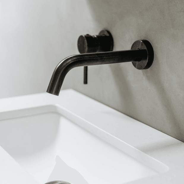 Close-up of a minimalist wall-mounted black metal bathroom tap with a curved spout above a white basin. The fitting is set against a smooth warm grey wall, with a clean, refined contemporary appearance.