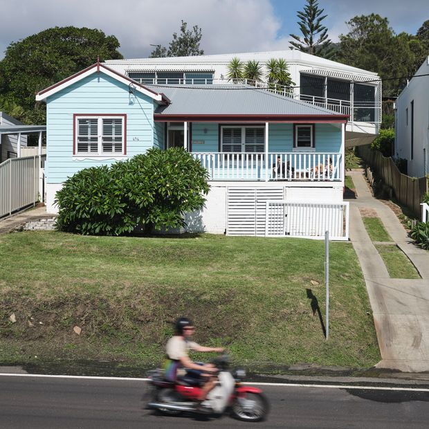 Street-facing view of a compact light-blue weatherboard house with white trim, a corrugated metal gabled roof, front verandah, raised garage/storage area, and a larger white upper-level structure behind it. The property sits on a grassy suburban slope beside a narrow concrete walkway and road.