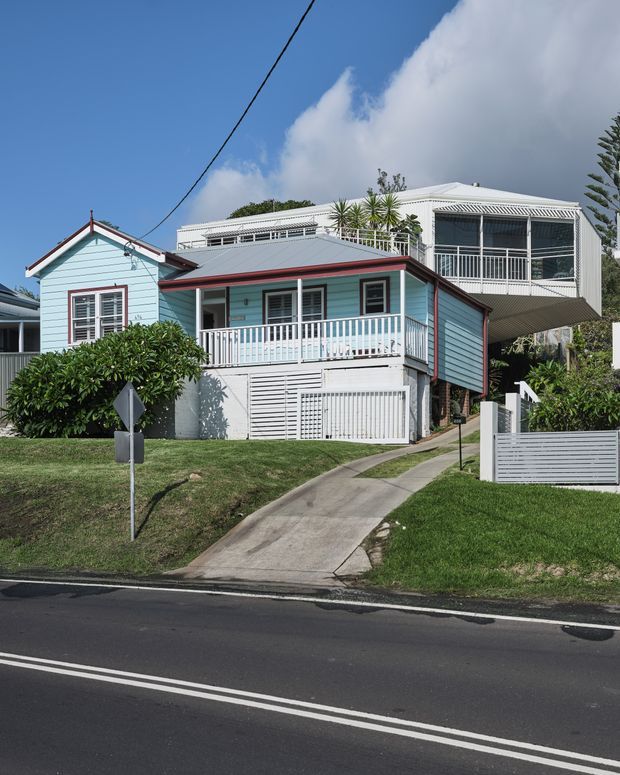 Street-facing view of a raised coastal-style timber house with pale blue weatherboard cladding, white trim, a red metal roof edge, front veranda and enclosed upper-level balcony. A concrete driveway leads to the elevated home, with a white slatted fence and suburban landscaping in front. A larger neighbouring house is visible behind.