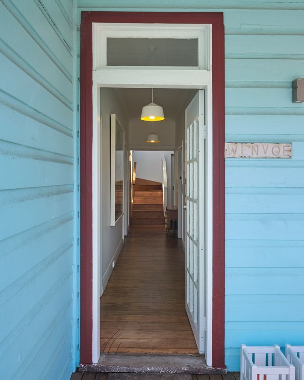 View from outside through an open entrance into a narrow residential hallway. The exterior is clad in bright turquoise painted horizontal weatherboards with a contrasting deep red door surround and white trim. Inside, the corridor has dark timber flooring, white walls, pendant lights, glazed doors, and a short flight of timber stairs leading upward. A small welcome sign is mounted beside the doorway.