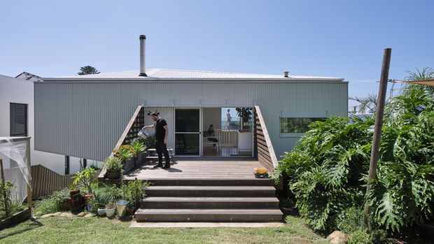 Exterior view of a raised, single-storey contemporary home with corrugated metal cladding and a low-pitched corrugated roof. A broad timber deck and stairway lead to glazed sliding doors, with lush planting around the entry. A chimney and utility poles are visible, and the image is photographed in bright daylight.