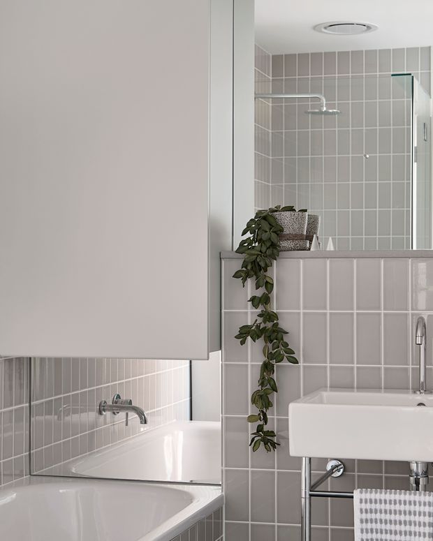 Bright, compact bathroom featuring pale grey tiled walls, a white bathtub, a wall-mounted white basin with exposed chrome plumbing, and a glass-enclosed shower. A large mirror reflects the bath area, while a trailing plant and woven accessories add softness to the clean tiled interior.