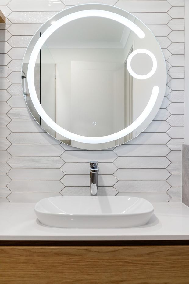 A bright, contemporary bathroom vanity featuring a round illuminated mirror, white vessel basin, chrome mixer tap, white engineered countertop, and warm timber cabinetry. The wall is finished with elongated light-toned hexagonal tiles.