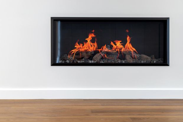 Minimalist interior detail showing a long, horizontal built-in linear fireplace with visible flames and dark stone or log media, set within a black surround on a smooth white wall above warm timber flooring.