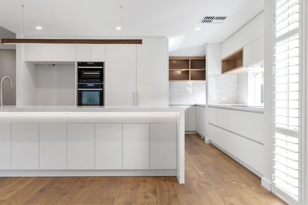 Bright contemporary kitchen with a large white island, handleless white cabinetry, integrated black appliances, pale stone surfaces, timber flooring, and a long horizontal timber-lined feature detail. A secondary galley-style kitchen zone with open timber shelving, window cabinetry, and white plantation shutters is visible to the right.