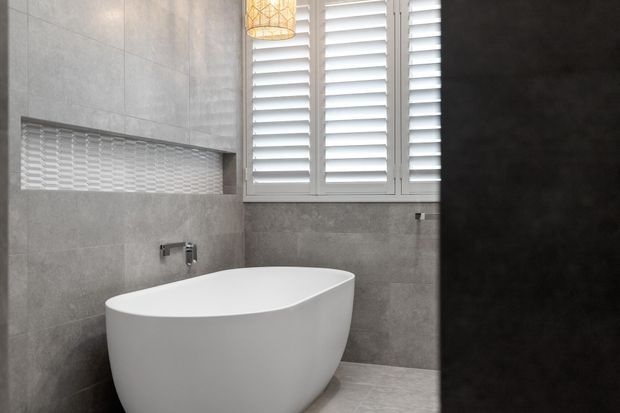 A contemporary bathroom featuring a freestanding white oval bathtub, large-format grey stone-look wall and floor tiles, a recessed tiled niche with textured white mosaic backing, and white plantation shutters. A warm pendant light adds a subtle decorative accent.