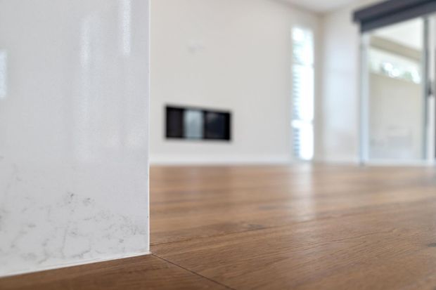 Close-up architectural interior view showing warm timber flooring extending through a bright, minimal space. A white stone or marble-clad vertical surface is prominent in the foreground, with a dark wall switch or outlet, white walls, window blinds, and dark-framed glazing visible in the softly blurred background.