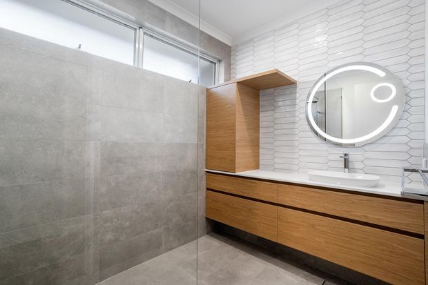 A bright contemporary bathroom featuring a floating timber vanity with integrated drawers, a white basin, and a round illuminated mirror. Large grey floor and wall tiles contrast with a white textured geometric tile splashback, while a frameless glass shower screen and high horizontal windows add openness and natural light.