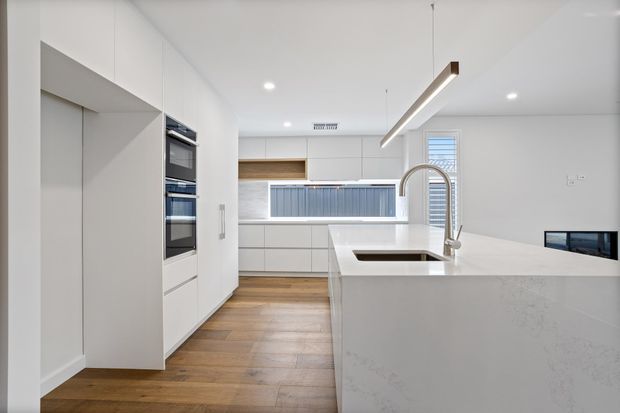 Bright, minimalist contemporary kitchen with extensive white flat-panel cabinetry, integrated double ovens, a large white island with a stainless-steel gooseneck tap and inset sink, and a long linear pendant light. Warm timber flooring contrasts with the predominantly white cabinetry and engineered-stone surfaces. A small glimpse of an adjacent living area is visible beyond the island.