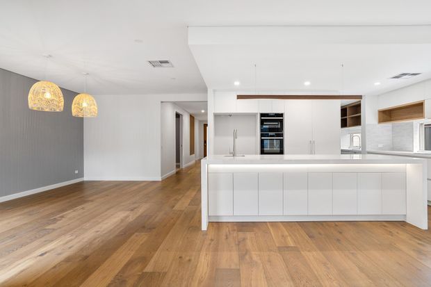 Bright contemporary open-plan kitchen and dining/living area with white handleless cabinetry, a large island with integrated seating, timber flooring, grey feature wall, and woven pendant lights. A hallway and additional rooms extend beyond the kitchen.