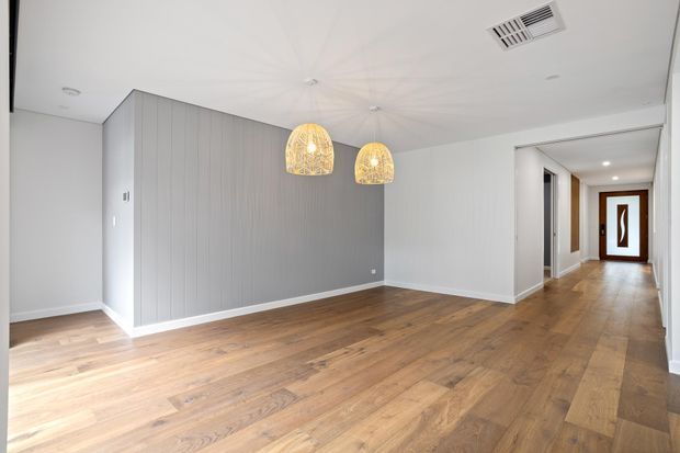A bright, empty open-plan interior with warm timber flooring, pale grey vertically panelled walls, white ceilings and trim, and two woven pendant lights. A hallway leads to a contemporary timber-framed front door with a decorative inset. The space appears suitable as a combined living and dining area.