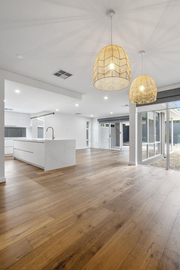 Bright open-plan kitchen and living area with a white island, integrated cabinetry, warm timber flooring, extensive glazing, and two prominent woven pendant lights. The room has white walls and ceiling, recessed lighting, and a clean contemporary fit-out.