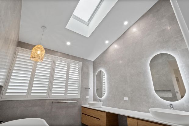 Bright contemporary bathroom with grey stone-look wall tiling, white double vanity basins, warm timber cabinetry, plantation shutters, a skylight, recessed lighting, and rounded illuminated mirrors. An arched opening adds a distinctive architectural feature.