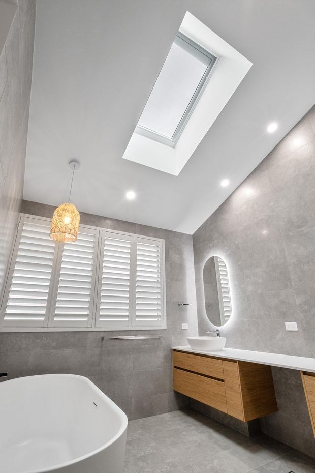 Bright contemporary bathroom featuring a freestanding white bathtub, floating timber vanity with white benchtop, vessel basin, illuminated oval mirror, grey tiled walls and floor, plantation shutters, recessed lighting, and a large skylight. A woven pendant adds warmth and texture.