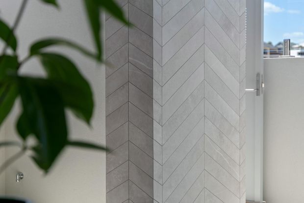 Close-up interior view of a pale grey chevron-pattern tiled feature wall beside a white-framed window, with a leafy plant partially obscuring the foreground. The image emphasizes the geometric wall finish and bright natural light.