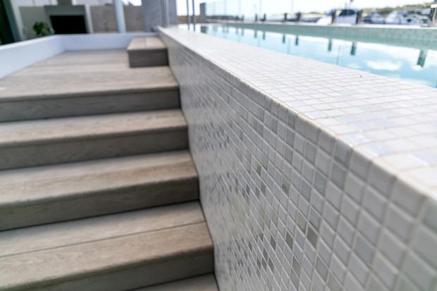 Close-up architectural detail of a raised swimming pool with small pale mosaic tile cladding, a tiled coping edge, and broad concrete steps leading up alongside the pool. Modern glazing and a bright outdoor setting are visible in the background.