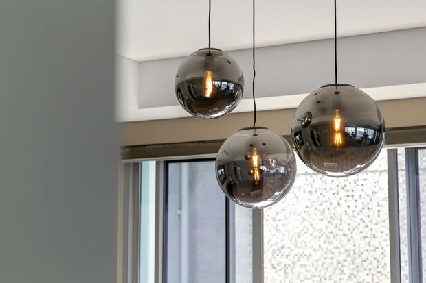 Close-up interior view of three smoked reflective glass globe pendant lights suspended at different heights from a recessed ceiling. Large windows and slim dark frames sit behind them, with a softly neutral contemporary architectural interior reflected in the glossy globes.