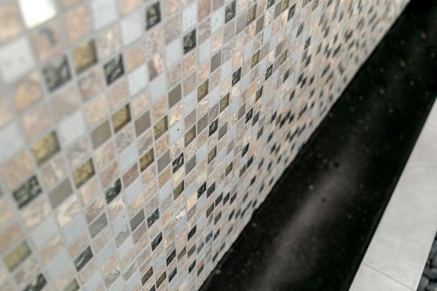 Close-up view of a wall finished with small square mosaic tiles in a varied neutral palette of beige, cream, grey, brown, and dark accents. A black stone or composite ledge runs along the base, with a small portion of adjacent light floor tile visible.
