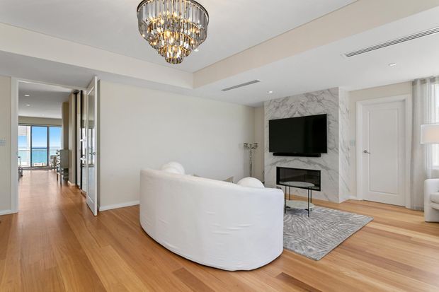 Bright, refined living room with pale walls, warm timber flooring, a curved white sofa, marble-look fireplace wall with wall-mounted television, patterned area rug, and a statement crystal chandelier. Open glazed doors reveal a bright adjoining space and distant water view.