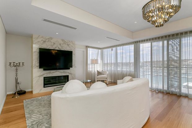 Elegant bright living room with floor-to-ceiling sheer-curtained glazing, a waterfront-facing outlook, white upholstered seating, light timber flooring, marble fireplace wall with mounted television, and a large crystal-and-metal chandelier. The room has a refined, luxurious contemporary fit-out with abundant natural light.