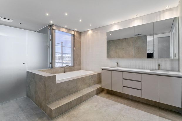 Contemporary bathroom with a freestanding-style built-in stone bathtub on a raised tiled platform, adjacent walk-in shower enclosure, and a long floating vanity with integrated basins and mirrored wall cabinetry. The room uses pale grey-beige stone finishes, ribbed wall detailing, recessed downlights, and a large window.