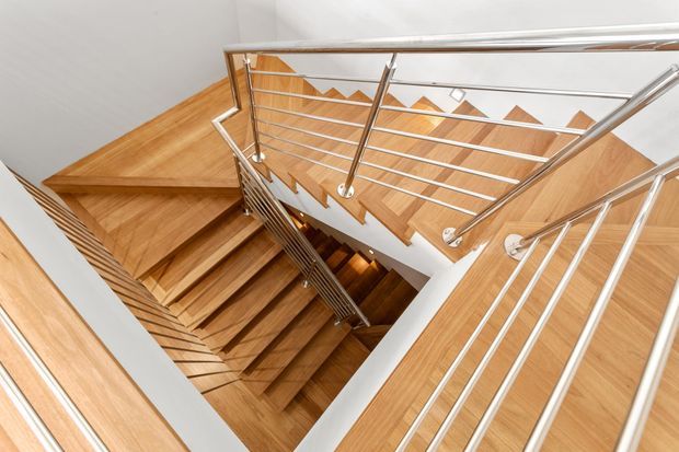 A bright multi-level stairwell viewed from above, featuring warm timber treads and landings, white walls, and contemporary stainless-steel horizontal cable-style balustrades. The stair flights form a dramatic open void with strong geometric lines and repeated angular detailing.