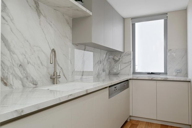 Compact contemporary kitchen with pale flat-panel cabinetry, a white marble-look stone benchtop and full-height matching splashback. The space includes an integrated dishwasher, undermount sink with brushed metal gooseneck tap, upper cabinets, and a window above the return bench. Warm timber flooring adds contrast to the predominantly white and grey finish palette.