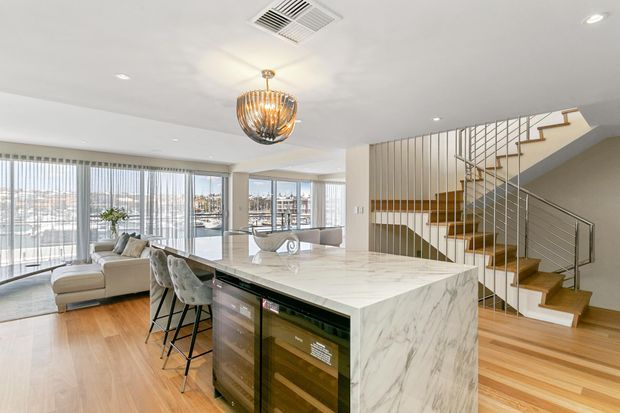 Bright open-plan apartment interior featuring a contemporary kitchen with a large white marble-look island, integrated wine storage, and bar seating. The space includes timber flooring, a sculptural pendant light, a living area, extensive floor-to-ceiling glazing, and a timber staircase with slim metal balustrades. An urban waterfront or marina-like view is visible beyond the windows.