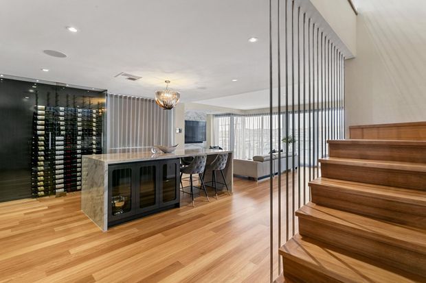 A refined open-plan residential interior featuring a dedicated wine display wall, a marble island or bar with dark built-in cabinetry, bar seating, a sculptural pendant light, and a timber staircase with slim vertical metal balustrade. Warm timber flooring and cabinetry contrast with white walls and extensive glazing.