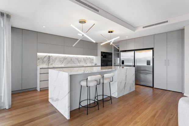 A bright contemporary kitchen with a large marble-look island and matching illuminated splashback, integrated grey cabinetry, stainless-steel French-door refrigerator, built-in appliances, timber flooring, and sculptural linear pendant lights. Two bar stools sit at the island, with sheer curtains and a neutral palette contributing to the refined modern interior.