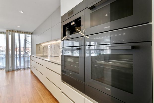 Contemporary galley-style kitchen with a long run of white handleless cabinetry, integrated stone benchtop and splashback, and a tall bank of dark glass Miele ovens and appliances. Warm timber flooring leads toward full-height glazed doors with sheer curtains, creating a bright, streamlined interior.