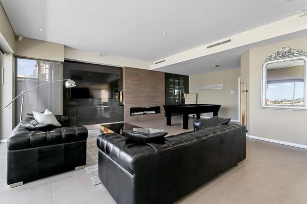A spacious contemporary living room with black leather sofas and armchair arranged around a low coffee table, facing a dark built-in media wall and television. A black grand piano sits in the adjoining area, while large windows, recessed ceiling lights, neutral walls, light flooring, and a decorative silver-framed mirror create a refined, open-plan interior.