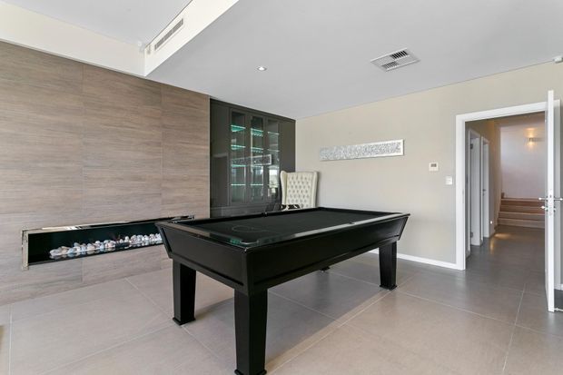 A contemporary recreation room featuring a black billiards table on large grey tiled flooring. The room has a full-height wood-look feature wall with a long recessed fireplace, dark built-in display cabinetry, neutral painted walls, recessed lighting, and a doorway leading to a short stairway.