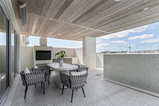 Contemporary covered rooftop terrace or balcony with an outdoor dining setting, built-in barbecue, pale terrazzo-style flooring, timber-look soffit, textured rendered walls, and a tiled privacy screen. Marina masts and water are visible beyond the terrace.