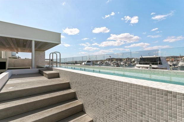 A bright rooftop or elevated terrace with a long tiled swimming pool overlooking a marina. Broad timber steps lead to the pool deck, while a minimalist flat-roofed pavilion provides shade at the left. The scene features crisp modern lines, pale neutral finishes, glass screening, and expansive views across boats and the waterfront.